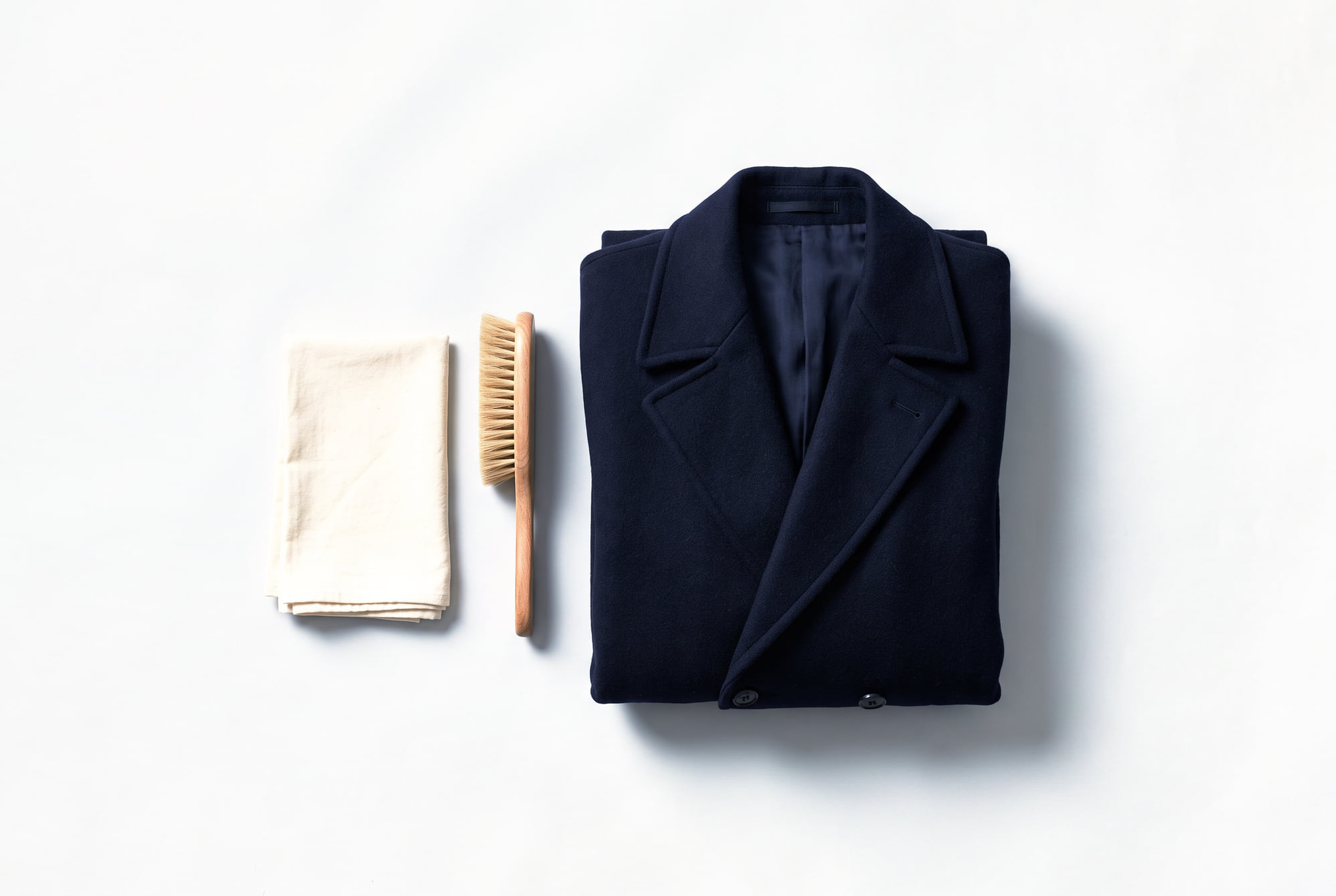 A folded navy wool overcoat beside a wooden clothes brush and a folded ivory cotton cloth.
